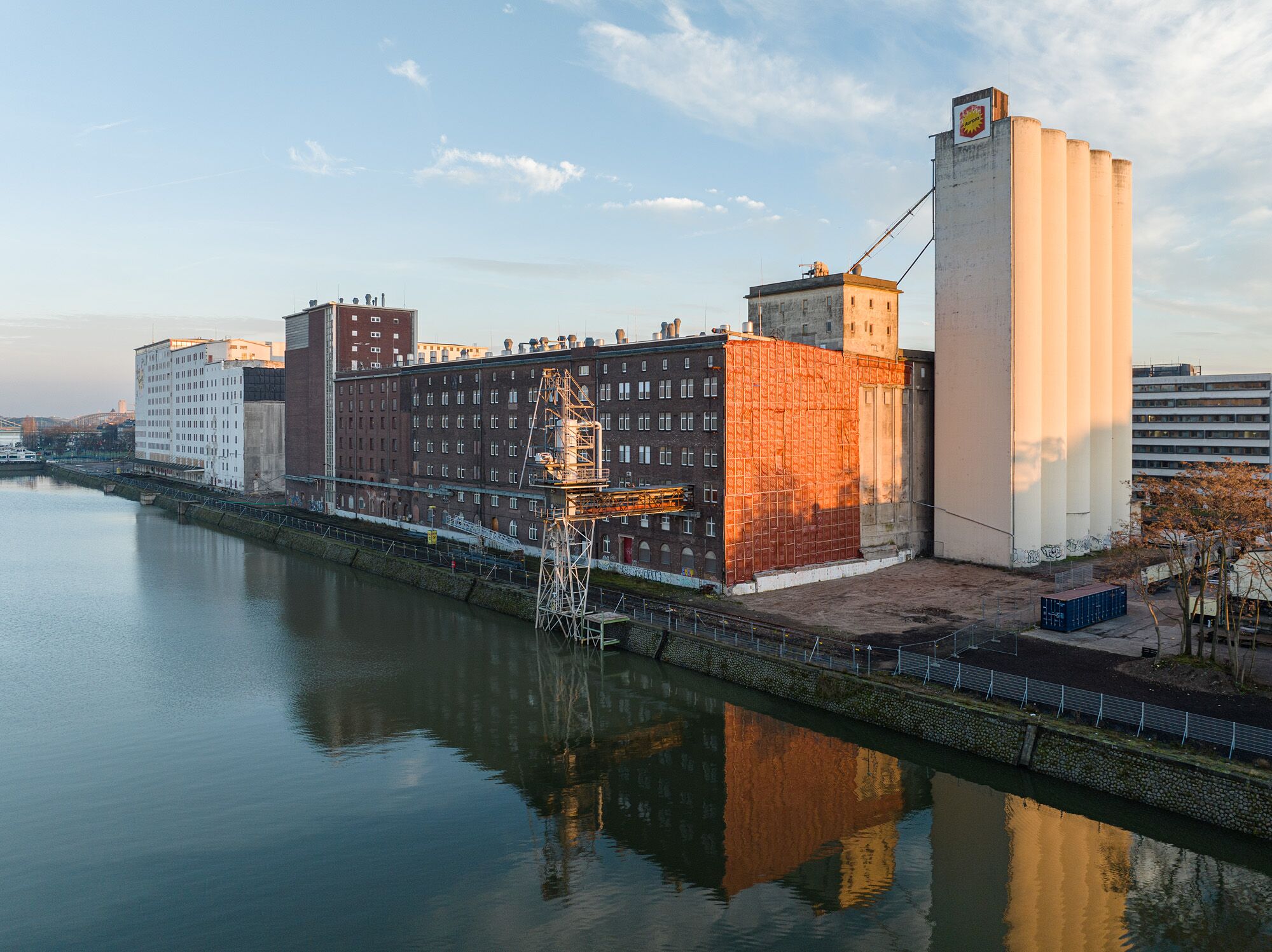 Ellmühle am Hafenbecken des Deutzer Hafens im Abendlicht: historische Backstein-Speichergebäude mit Verladekran an der Kaimauer und die hohen Silotürme mit Aurora-Schild, die sich im ruhigen Wasser spiegeln